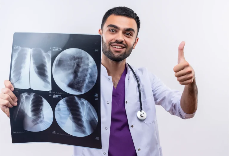 Pleased young male doctor wearing stethoscope medical gown holding x ray his thumb up isolated white background